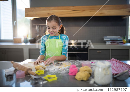 Child girl wearing apron pressing cookie cutter into dough on countertop in kitchen by window Child girl wearing apron pressing cookie cutter into dough on countertop in kitchen by window 132987397