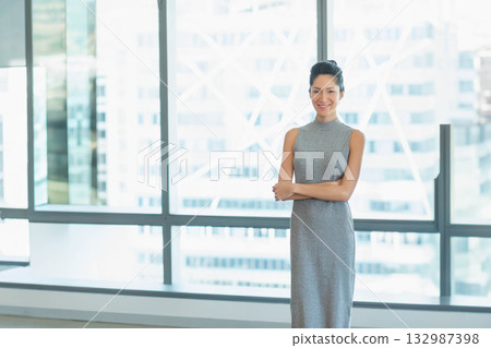 Asian woman in fitted grey dress crossing arms by floor-to-ceiling windows overlooking skyline 132987398
