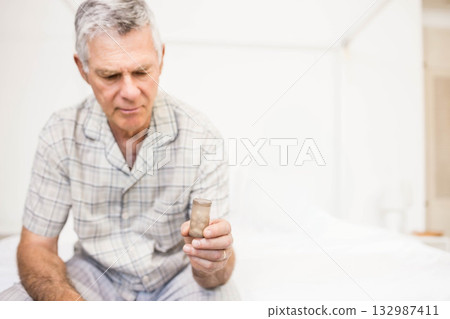 Senior man wearing checkered pajamas holding amber pill bottle and sitting on white bed in bedroom 132987411