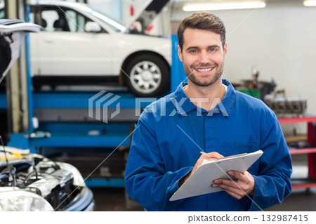 Male mechanic wearing blue coveralls standing in repair bay holding clipboard and pen, copy space 132987415
