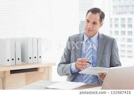 Man wearing gray suit striped tie holding clipboard reviewing documents at office desk, copy space Man wearing gray suit striped tie holding clipboard reviewing documents at office desk, copy space 132987418