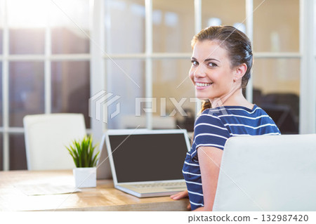 Woman using laptop and smiling at camera at office meeting table with scattered papers, copy space Woman using laptop and smiling at camera at office meeting table with scattered papers, copy space 132987420