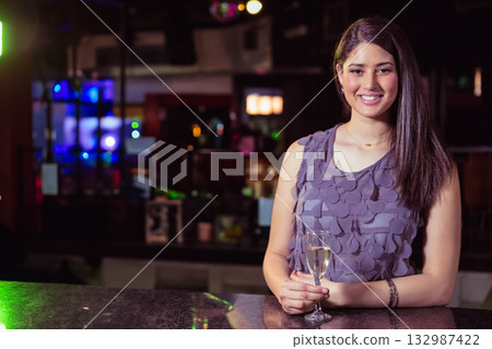 Woman in her twenties holding champagne flute behind bar counter at bar lounge, copy space 132987422