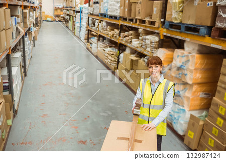 Female warehouse worker applying packing tape onto box in warehouse aisle with shelving, copy space Female warehouse worker applying packing tape onto box in warehouse aisle with shelving, copy space 132987424