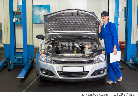 Female auto tech with clipboard inspecting engine under hood open on silver sedan in repair bay 132987425