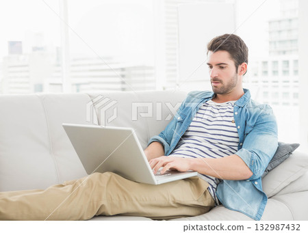 Man reclining on leather sofa typing on laptop in apartment living room with pillow, copy space 132987432