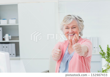 Senior woman raising both arms giving thumbs-up gesture sitting at white desk with monitor 132987433