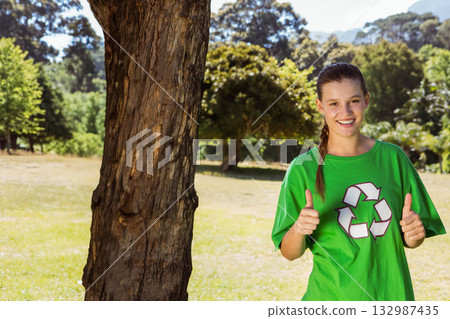Woman standing next to tree trunk in sunny meadow wearing green recycling shirt, copy space Woman standing next to tree trunk in sunny meadow wearing green recycling shirt, copy space 132987435