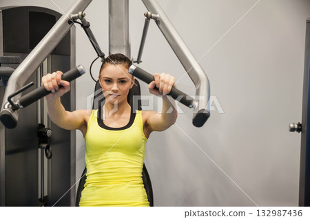 Woman wearing yellow tank top pushing padded handles forward on chest fly machine at fitness center Woman wearing yellow tank top pushing padded handles forward on chest fly machine at fitness center 132987436