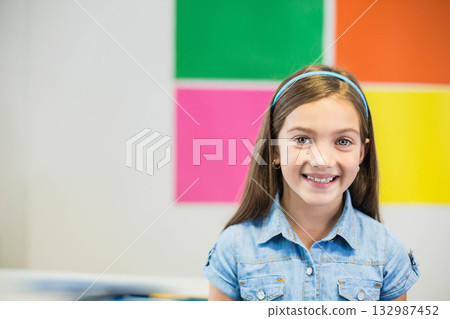 Girl wearing denim shirt, blue headband and hoop earrings smiling in classroom with colorful panels Girl wearing denim shirt, blue headband and hoop earrings smiling in classroom with colorful panels 132987452