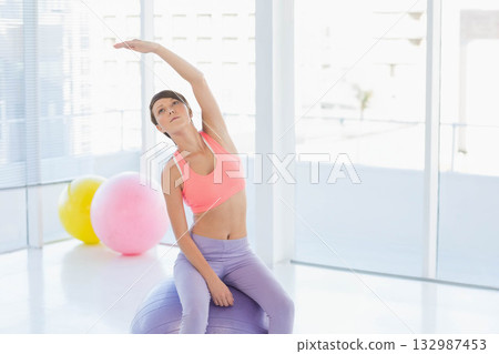 Woman in sportswear balancing on purple exercise ball stretching side by windows at fitness studio Woman in sportswear balancing on purple exercise ball stretching side by windows at fitness studio 132987453