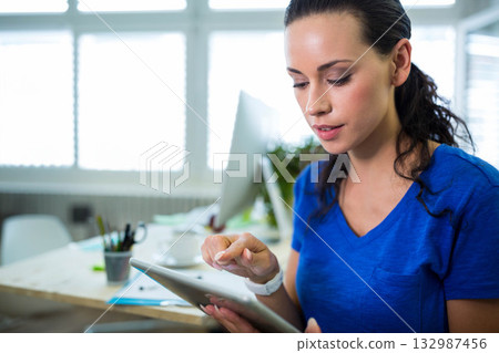 Woman tapping tablet sitting at desk in office with monitor and coffee cup, copy space 132987456