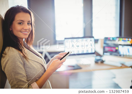 Woman in gray blazer leaning on wall at office with smartphone and two monitors, copy space Woman in gray blazer leaning on wall at office with smartphone and two monitors, copy space 132987459