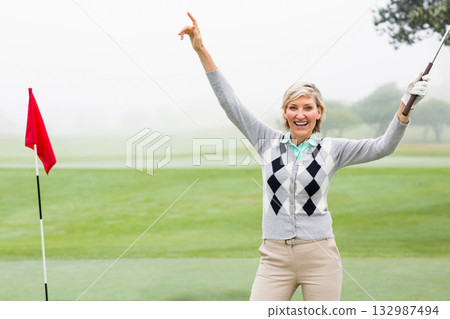 Senior golfer standing on golf green by red flagstick holding club, pointing skyward, copy space 132987494