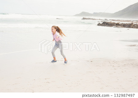 Girl running along wet sand at water's edge on beach wearing pink sportswear and sneakers 132987497
