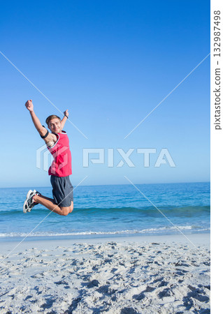 Man jumping on sandy shoreline wearing red top black armband white sneakers, copy space Man jumping on sandy shoreline wearing red top black armband white sneakers, copy space 132987498