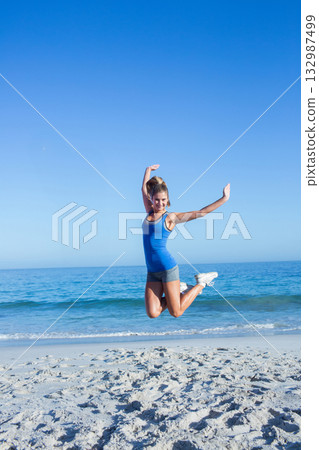 Woman leaping joyfully above wet sand on calm shoreline wearing tank top, shorts, running shoes Woman leaping joyfully above wet sand on calm shoreline wearing tank top, shorts, running shoes 132987499