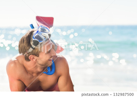 Male snorkeler leaning on shoreline wearing red trunks with snorkel gear and dive fins, copy space 132987500