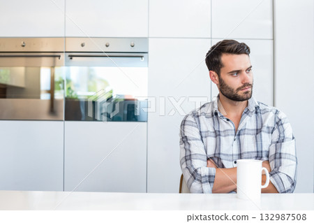 Male in late twenties sitting at modern kitchen countertop holding mug facing ovens, copy space 132987508