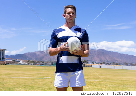 Male athlete wearing striped jersey and white shorts standing on sports field holding rugby ball 132987515