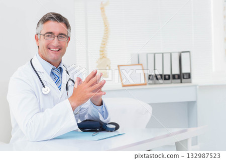 Smiling male physician wearing lab coat at desk in clinic with stethoscope, spine model, copy space Smiling male physician wearing lab coat at desk in clinic with stethoscope, spine model, copy space 132987523