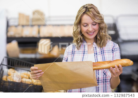 Female customer selecting fresh baguette and packing into brown paper bag at bakery display Female customer selecting fresh baguette and packing into brown paper bag at bakery display 132987543