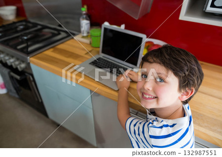 School-aged boy standing at wooden kitchen counter typing on laptop beside stove, copy space School-aged boy standing at wooden kitchen counter typing on laptop beside stove, copy space 132987551