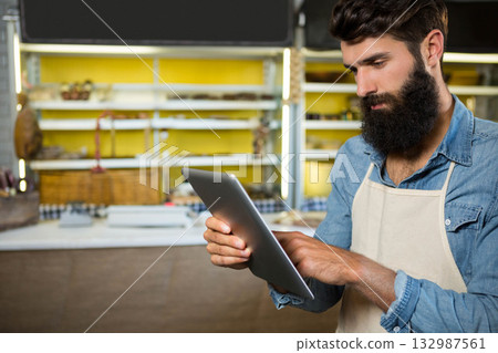 Bearded man wearing apron using tablet behind bakery counter, scale and bread baskets copy space Bearded man wearing apron using tablet behind bakery counter, scale and bread baskets copy space 132987561