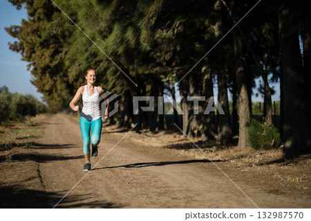 Female runner jogging along forest-edge dirt trail in white sleeveless top and turquoise leggings Female runner jogging along forest-edge dirt trail in white sleeveless top and turquoise leggings 132987570