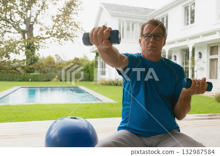 Senior White male in sportswear lifting blue dumbbells on pool deck with exercise ball, copy space 132987584