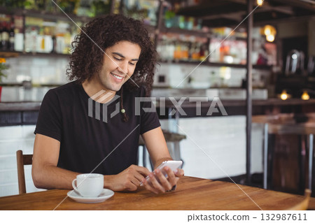 Man sitting at wooden table in cafe smiling at smartphone with ceramic coffee cup and saucer 132987611
