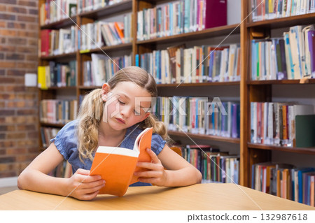 Female child reading orange-covered paperback at library table near brick wall, bookshelves Female child reading orange-covered paperback at library table near brick wall, bookshelves 132987618