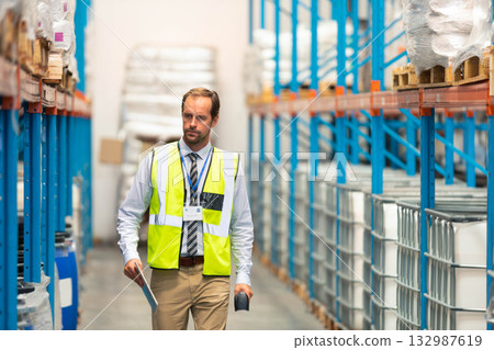 Man wearing safety vest and ID badge walking through warehouse aisle holding scanner and folder Man wearing safety vest and ID badge walking through warehouse aisle holding scanner and folder 132987619