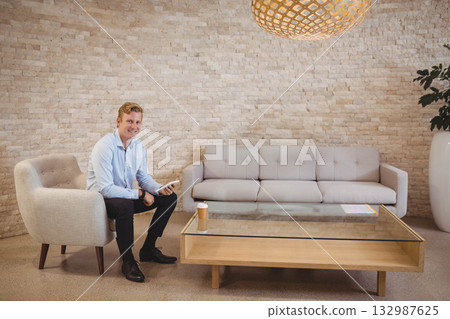 Man sitting on armchair in office lounge holding tablet and coffee cup beside planter, copy space 132987625