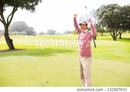 Asian mid adult man celebrating putt on golf green holding putter and flagstick, copy space 132987633