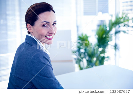 Businesswoman in suit smiling at camera, turning at office desk with horizontal blinds and plant 132987637