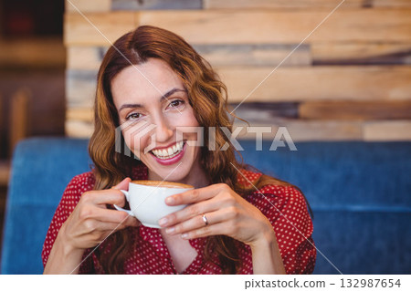 Middle-aged woman in red blouse smiling and holding latte on blue bench in coffee shop Middle-aged woman in red blouse smiling and holding latte on blue bench in coffee shop 132987654
