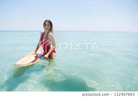 Female surfer in red swimsuit holding surfboard standing in turquoise sea water under sunny sky 132987665