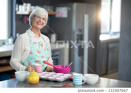 Senior woman pouring cupcake batter into muffin tin on countertop wearing floral apron, copy space Senior woman pouring cupcake batter into muffin tin on countertop wearing floral apron, copy space 132987667