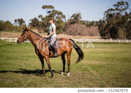 Woman riding brown horse around grass paddock near white fence wearing helmet and boots Woman riding brown horse around grass paddock near white fence wearing helmet and boots 132987670