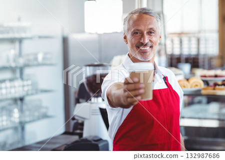 Middle-aged barista holding paper coffee cup and smiling by pastry display case in coffee shop 132987686