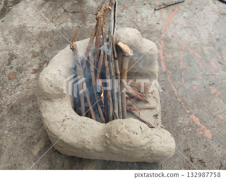 Traditional Indian mud chulha with fire burning inside using wooden logs, showcasing rural cooking method with glowing flames and smoke during winter or daily meal preparation in village households 132987758