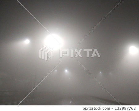 Foggy night scene with glowing streetlights illuminating the misty road, creating a calm and atmospheric view of the street under thick haze during nighttime Foggy night scene with glowing streetlights illuminating the misty road, creating a calm and atmospheric view of the street under thick haze during nighttime 132987768