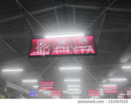 Railway station interior with no smoking, no spitting, and prohibited items signboards in Hindi and English under metal roof with fluorescent lights and light fog, India public notice signage 132987810