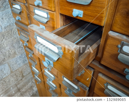 Vintage wooden drawer cabinet with metal handles and label holders, open empty drawer in old library card catalog filing system, retro office furniture storage unit with rustic archive organization Vintage wooden drawer cabinet with metal handles and label holders, open empty drawer in old library card catalog filing system, retro office furniture storage unit with rustic archive organization 132989085
