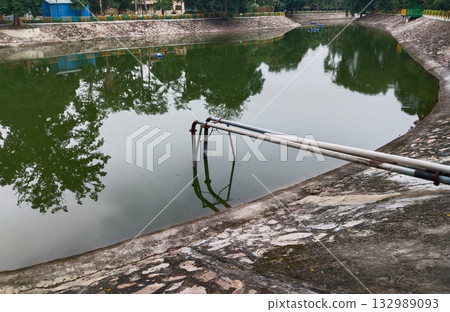 Water canal pond with stone embankment and metal pipes, outdoor artificial reservoir in public park used for irrigation and water storage surrounded by greenery and reflecting trees in calm surface 132989093