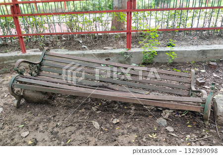 Old rusty iron bench with decorative armrest, lying askew on muddy ground with a tree branch, public park environment, neglected condition Old rusty iron bench with decorative armrest, lying askew on muddy ground with a tree branch, public park environment, neglected condition 132989098