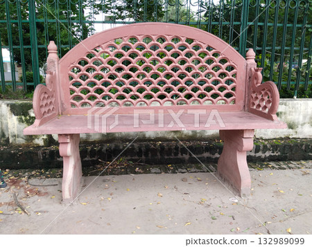 Ornate pink stone bench with traditional lattice-pattern backrest and decorative armrests, installed outdoors in park setting, blending heritage architecture with public seating for relaxation Ornate pink stone bench with traditional lattice-pattern backrest and decorative armrests, installed outdoors in park setting, blending heritage architecture with public seating for relaxation 132989099