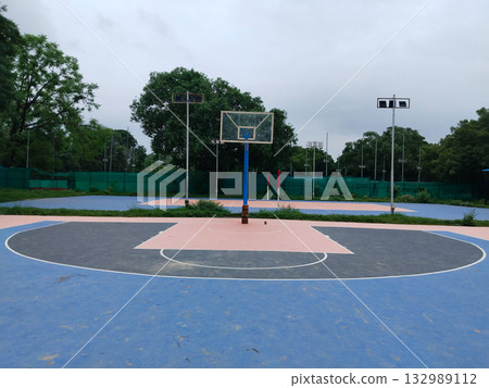 Empty outdoor basketball court with pink and blue painted surface, lone hoop post in quiet sports complex surrounded by greenery and tall light poles, peaceful athletic ground for games and recreation Empty outdoor basketball court with pink and blue painted surface, lone hoop post in quiet sports complex surrounded by greenery and tall light poles, peaceful athletic ground for games and recreation 132989112