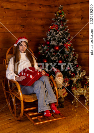A girl in a Santa hat sits in a rocking chair surrounded by a Christmas tree A girl in a Santa hat sits in a rocking chair surrounded by a Christmas tree 132989290